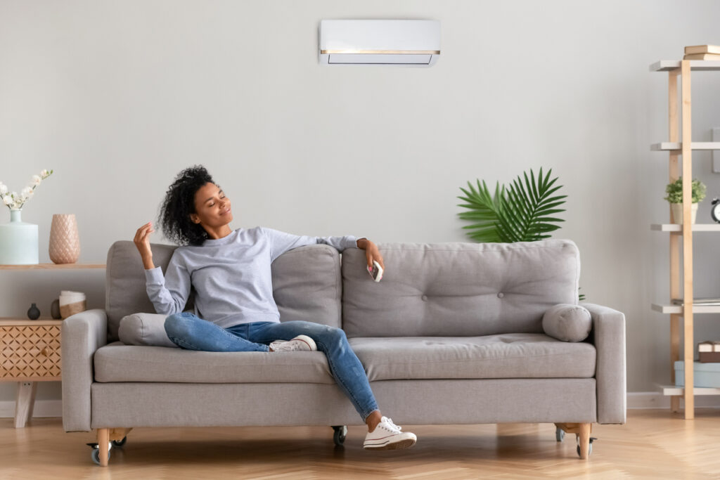 Woman sitting on couch relaxing; ductless mini-split on the wall in the background blowing cool air.